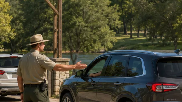 A park ranger directs an orderly line of cars through a green riverside park entrance.